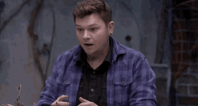 a man in a purple plaid shirt is sitting at a table in a dark room with his mouth open .