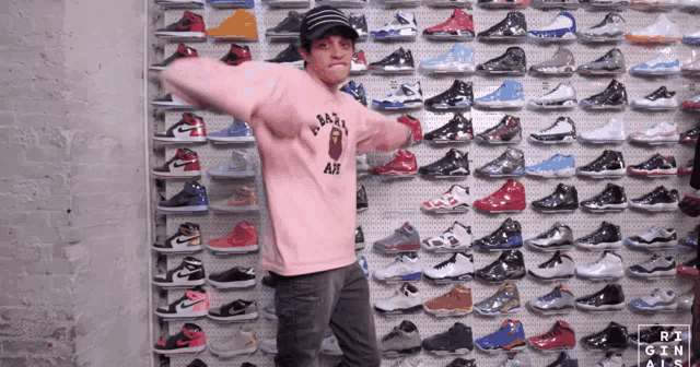 a man wearing a pink bape sweatshirt stands in front of a wall of sneakers