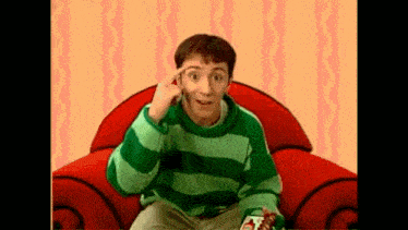 a man in a green and blue striped sweater is sitting in a red chair with his hand on his head .