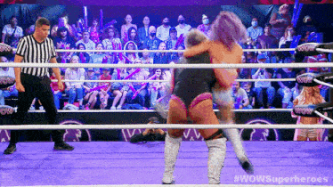 two women are wrestling in a ring with a referee in front of a crowd that is watching .