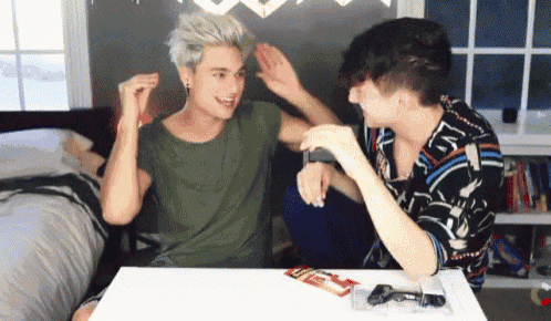 two young men are sitting at a table playing a video game and one of them is wearing a green shirt and the other is black