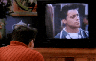 a man in an orange sweater is sitting in front of a television watching a movie .