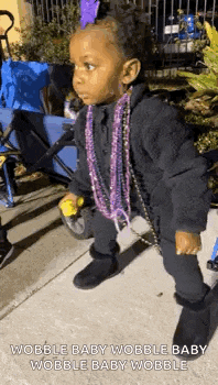 a little girl wearing a purple necklace and a black jacket is standing on the sidewalk .