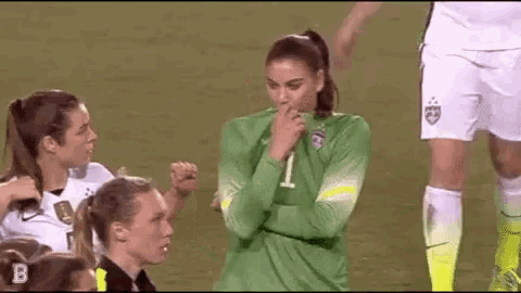 a female soccer player is covering her mouth with her hand while standing on a field with other players .