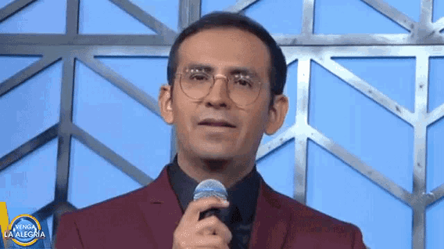 a man wearing glasses and a red suit holds a microphone in front of a viva la alegria logo