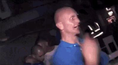 a bald man in a blue shirt is dancing in a dark room with a group of people .