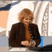 a woman in a brown jacket is sitting at a desk holding a small object in her hands .