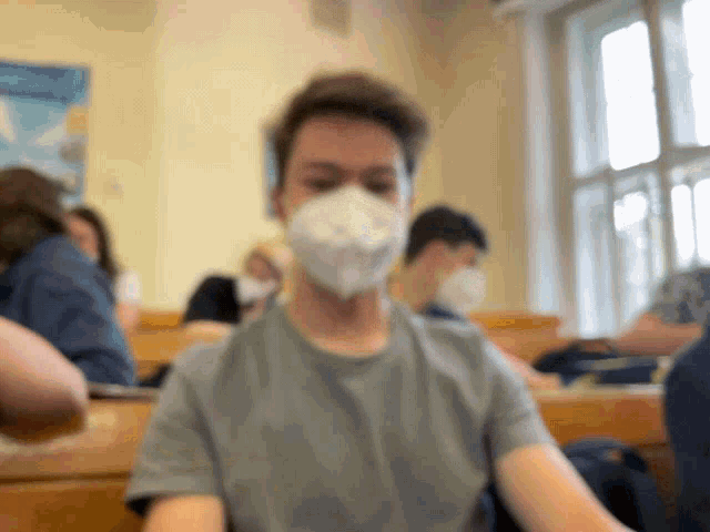 a man wearing a mask sits in a classroom with other people