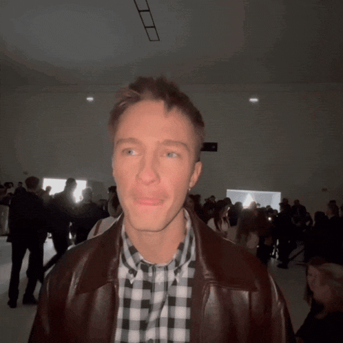 a man wearing a plaid shirt and a brown jacket stands in a crowded room