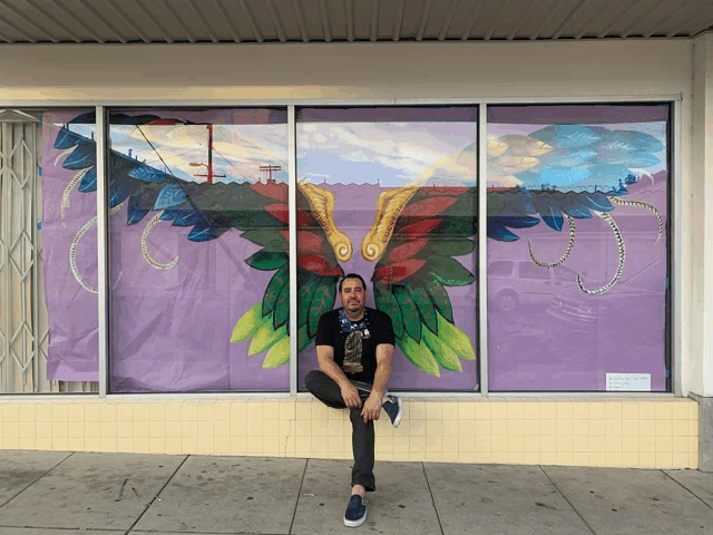 a man sits on the sidewalk in front of a window with a painting of wings on it