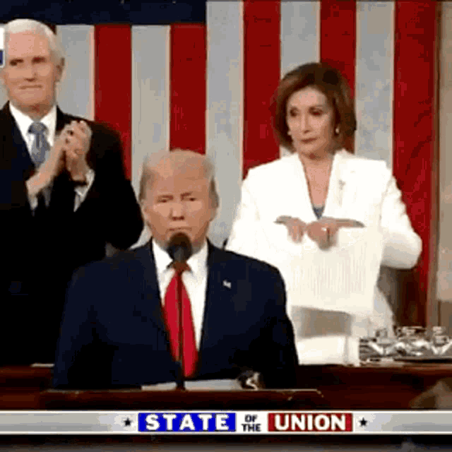 a man in a suit and tie is speaking into a microphone while a woman in a white jacket stands behind him