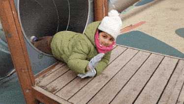 a girl in a green jacket and white hat is laying on a wooden deck