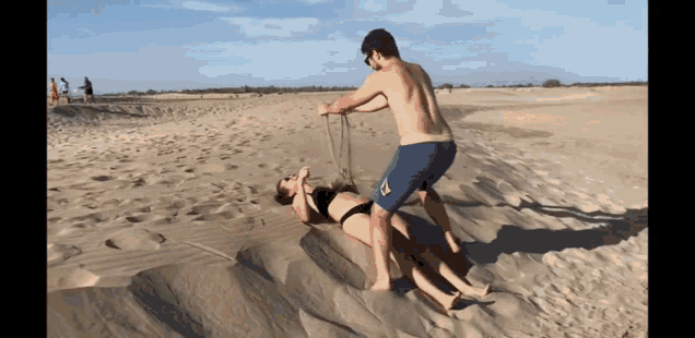 a man in blue shorts is pulling a woman in a bikini up a sand dune