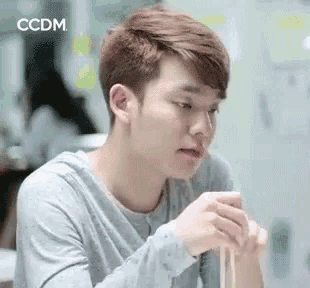 a young man is sitting at a table eating noodles with chopsticks and looking at his phone .