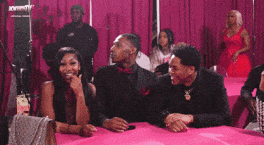a group of people sitting at a table with a pink curtain behind them that says now thatstv