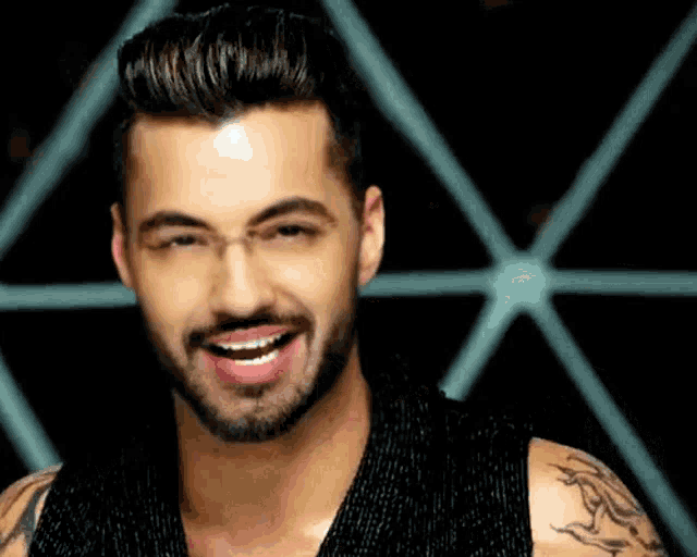 a man with a beard and tattoos is smiling in front of a geometric background .