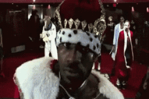 a man wearing a crown and a fur collar is standing on a red carpet .