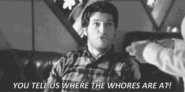 a man in a plaid shirt is sitting on a couch talking to another man who is pointing at him