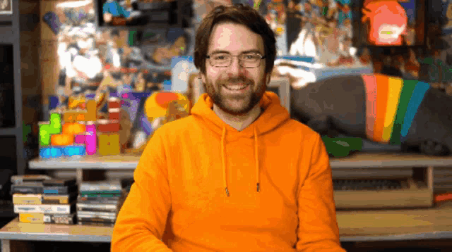 a man wearing glasses and an orange hoodie is smiling in front of a stack of games