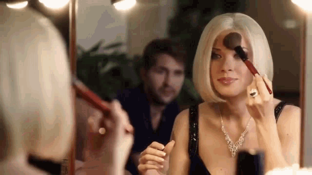 a woman applying makeup in front of a mirror with a man in the background