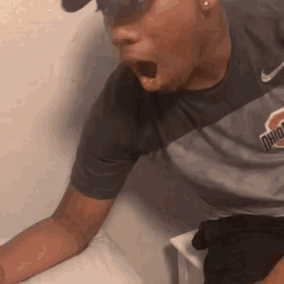 a man wearing sunglasses and a ohio shirt is sitting on a bed with his mouth open .