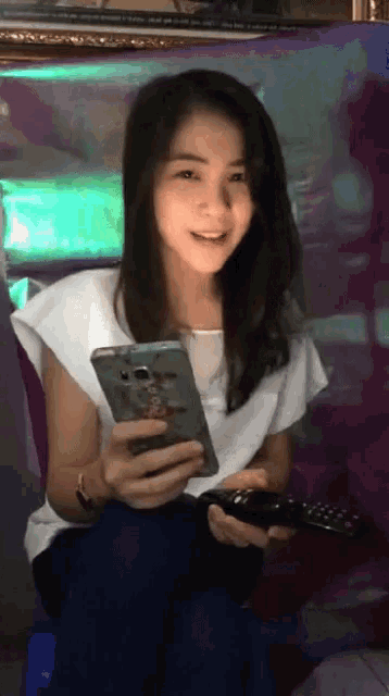 a woman in a white shirt is holding a cell phone and a remote