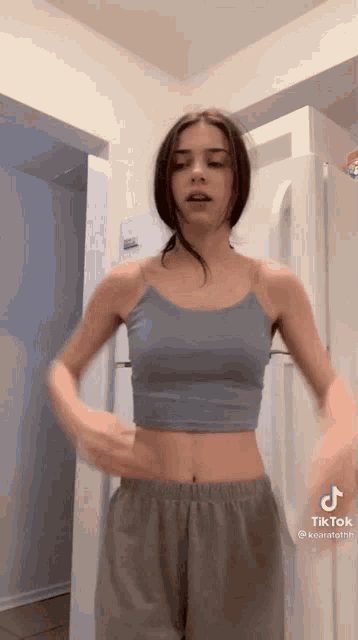 a woman wearing a blue crop top and grey pants is standing in front of a refrigerator .