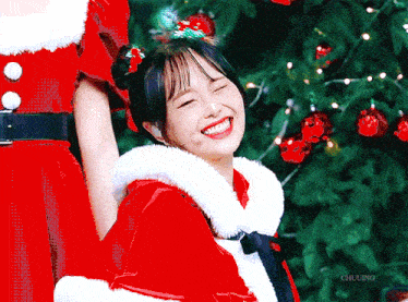 a girl in a santa outfit is smiling with a christmas tree in the background