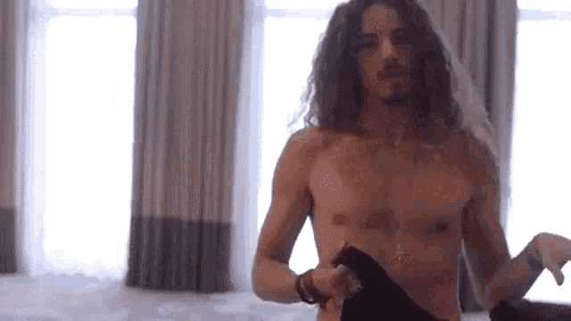 a shirtless man with long hair and a beard is standing in front of a window .