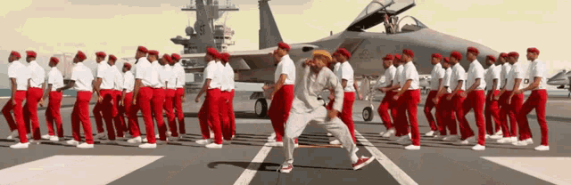 a group of men in red pants are dancing in front of an airplane
