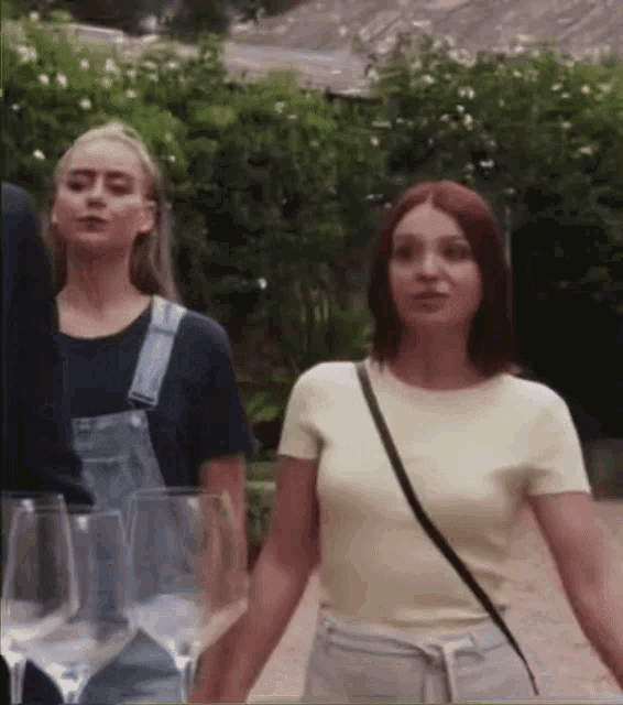 two women are holding hands while walking down a path with wine glasses in the background