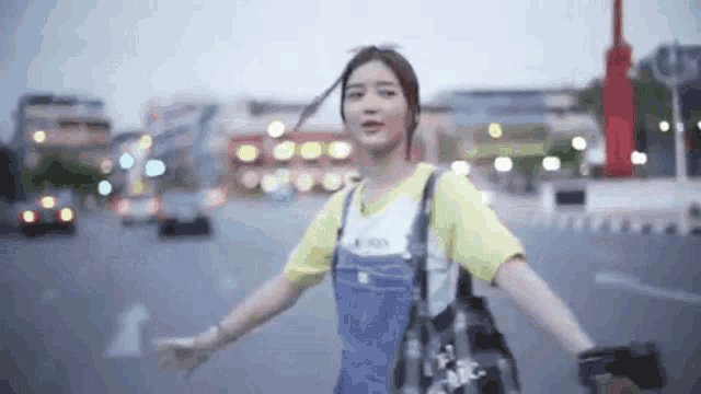 a woman wearing overalls and a yellow shirt is standing on a street with her arms outstretched
