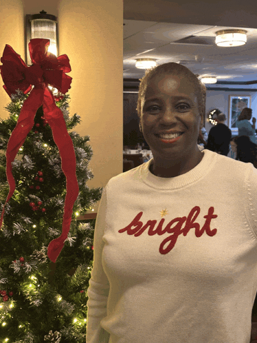 a woman wearing a white sweater that says bright is smiling in front of a christmas tree