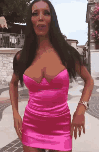 a woman in a pink dress is standing on a sidewalk and looking at the camera .