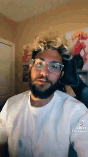a man with a beard wearing glasses and a white shirt is sitting in a chair .