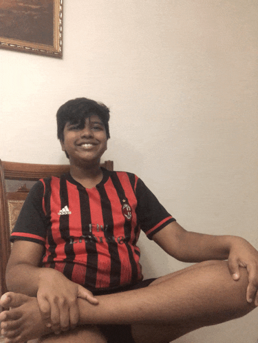 a boy wearing a red and black adidas shirt sits on a chair with his legs crossed