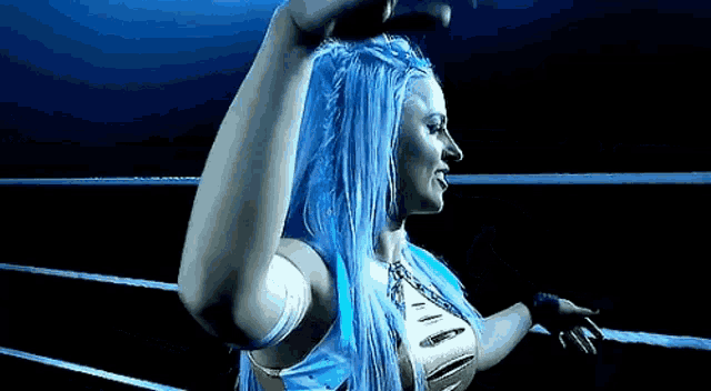 a woman with blue hair is standing in a wrestling ring with her arms up .