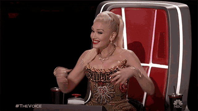 a woman in a gold dress is sitting in a chair with a cup that says nbc on it