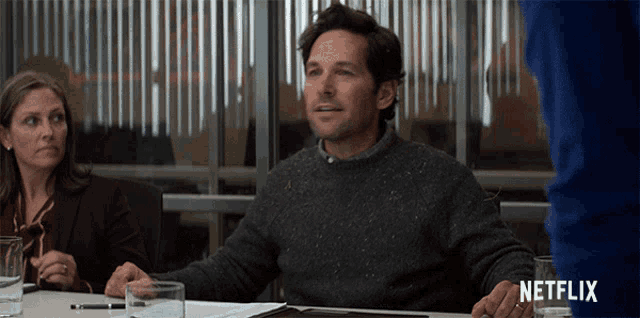 a man in a grey sweater sits at a table with a netflix logo behind him