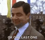 mr bean is wearing a suit and tie and smiling while saying `` ok one last one '' .