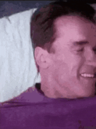 a man in a purple shirt is laying on a bed with a white pillow .