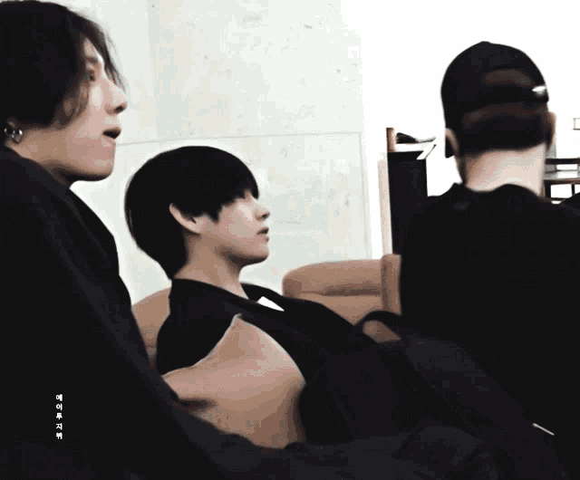 three men are sitting in a room and one of them is wearing a baseball cap