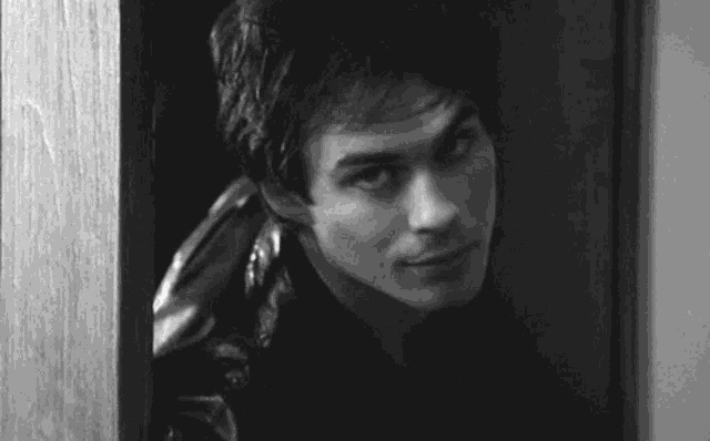 a black and white photo of a man peeking out of a door .