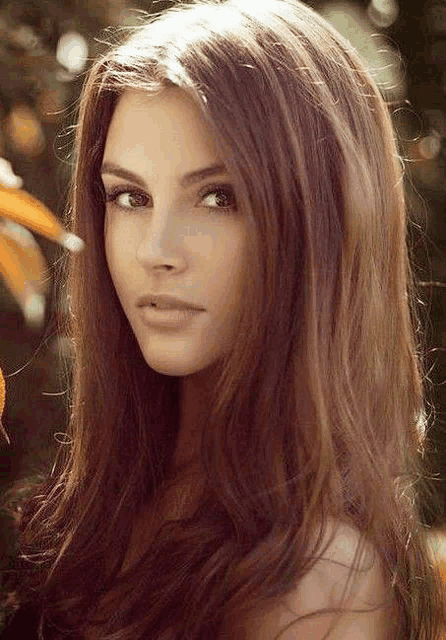 a close up of a woman 's face with long brown hair and a flower in the background .