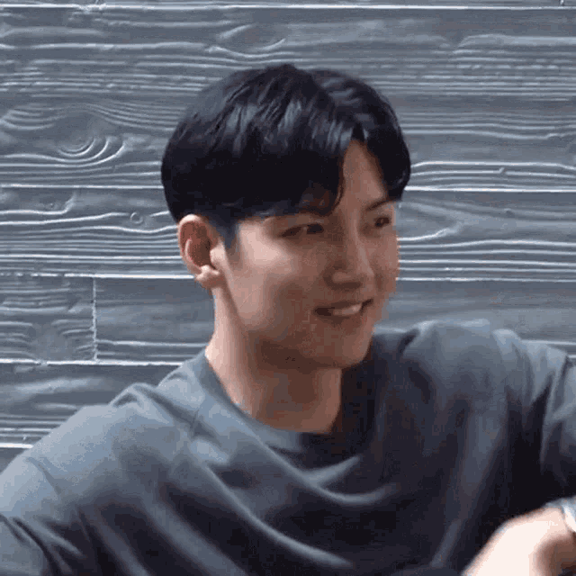a young man wearing a grey shirt is smiling and looking at the camera while sitting in front of a wooden wall .
