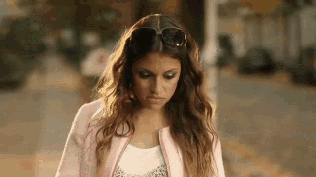 a woman wearing sunglasses and a pink jacket looks down at something