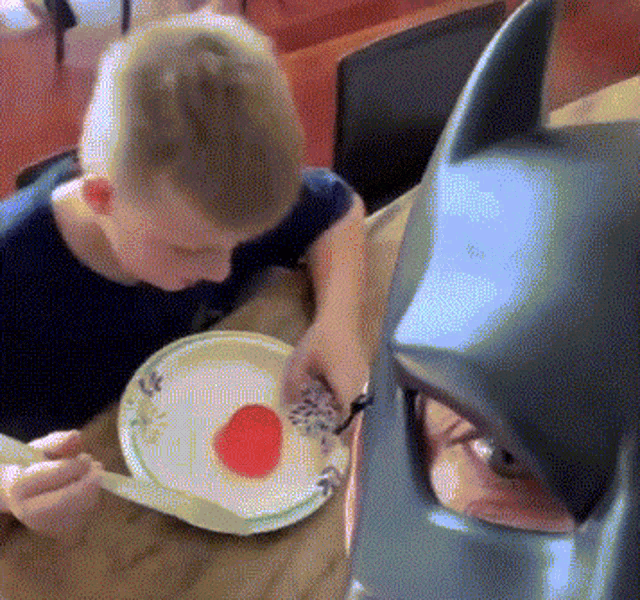 a boy is eating a plate of food next to a batman mask .