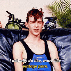 a man in a blue tank top is sitting on a couch and says i actually do like more like vintage porn