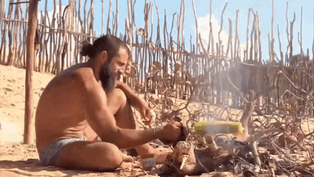 a man without a shirt is sitting on the beach cooking corn on the cob .