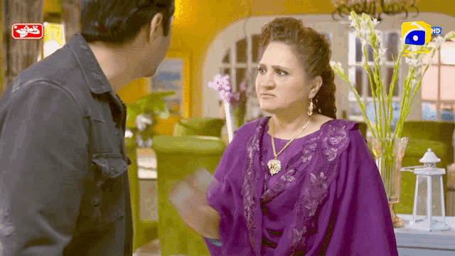 a woman in a purple dress is talking to a man in a black jacket with a g logo on the bottom right